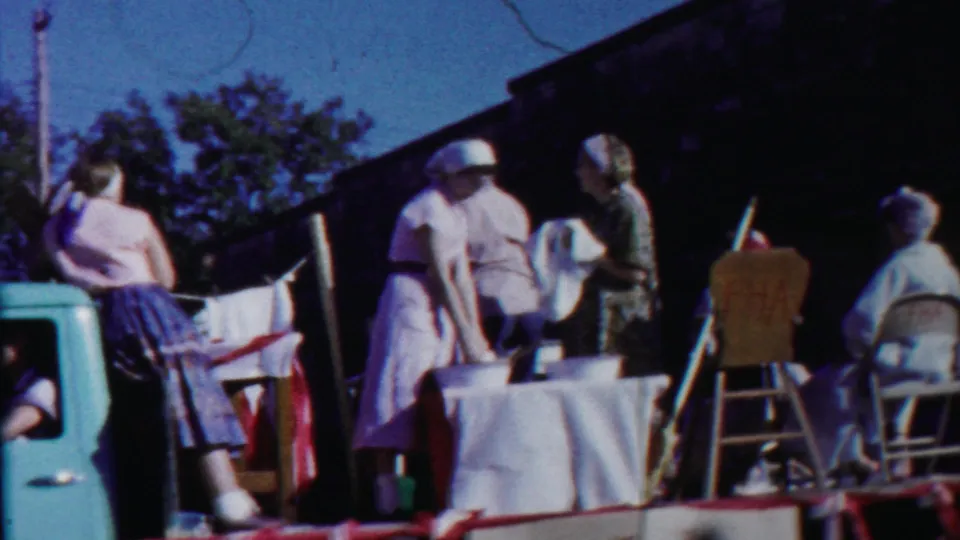 Women on a 1958 Cheyenne County parade float stand near a table and chairs, with trees and a building in the background.