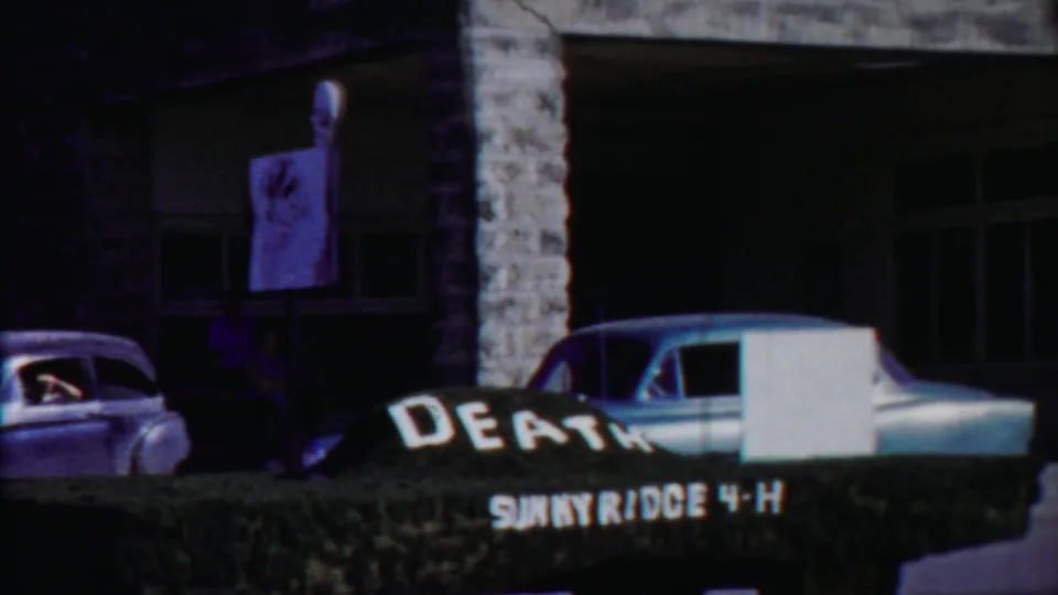 End of the 1958 Cheyenne County parade view with a hand-painted 4-H sign, vintage car, and stone building visible.
