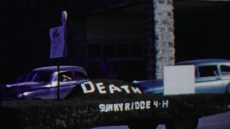 1958 Cheyenne County, Kansas home-movie frame showing a dark sedan near a hedge sign reading DEATH SUNNYRIDGE 4-H.