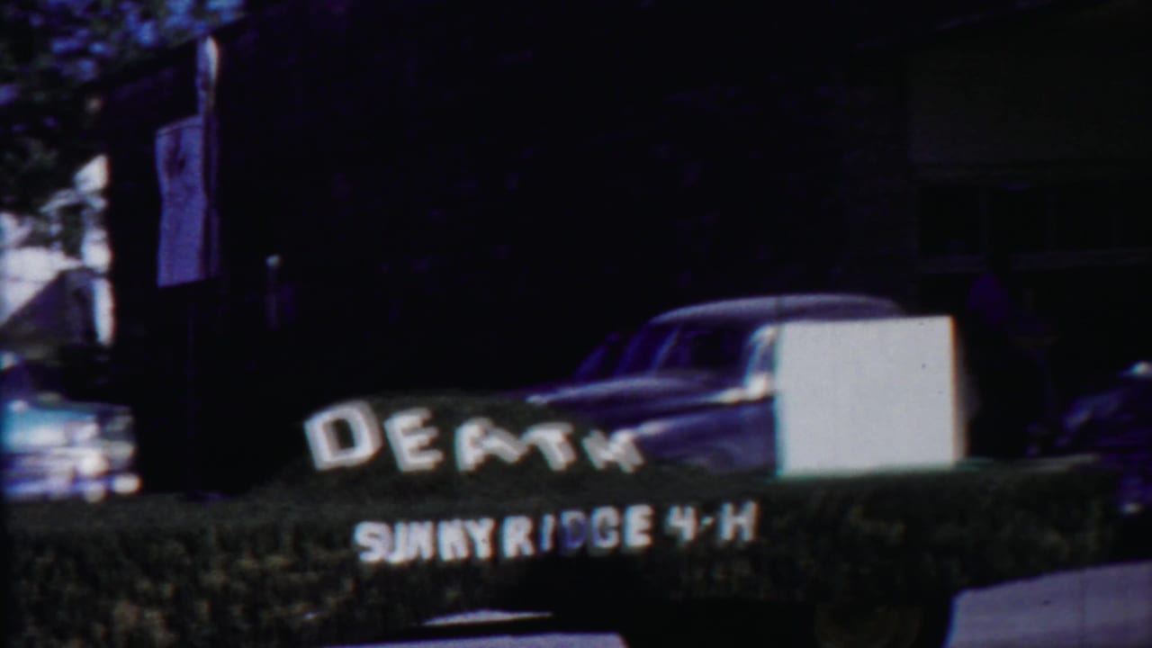 Blue pickup and nearby parked car pass a Sunnyridge 4-H hedge sign with DEATH lettering in the 1958 Kansas footage.