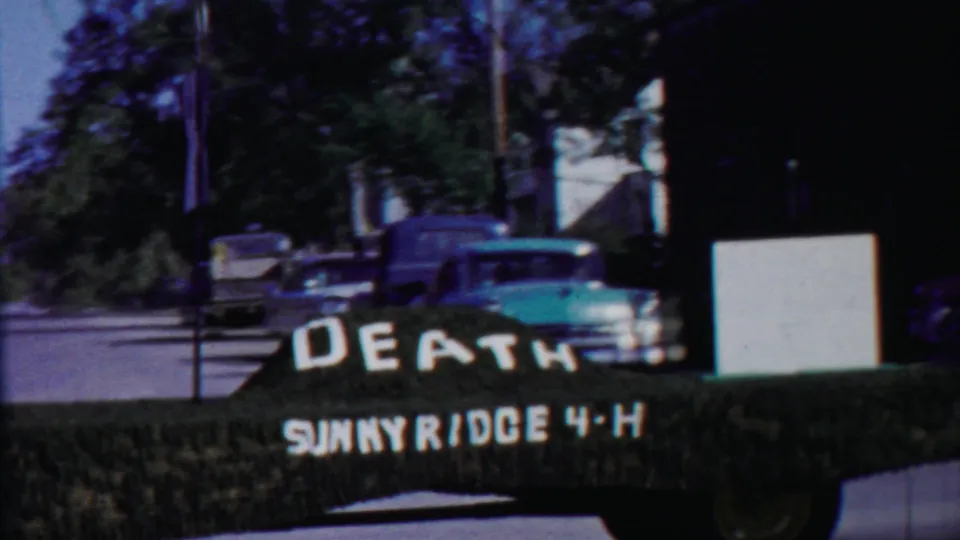 Roadside home-movie view in Cheyenne County, Kansas, with a pickup, street signs, trees, and the DEATH SUNNYRIDGE 4-H display.
