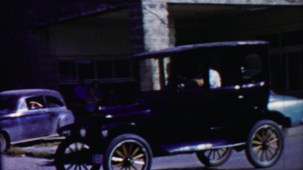 Black vintage car passing a stone building with a parked sedan nearby.