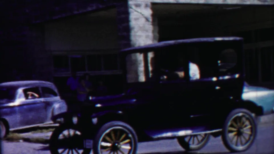 A black Model T-style car drives through a Cheyenne County, Kansas street scene with a mid-century sedan and stone building visible.