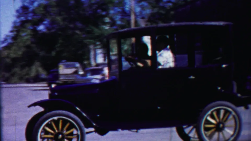 A black Model T-style car moves along the street, framed against trees and parked mid-century cars in the Kansas town setting.