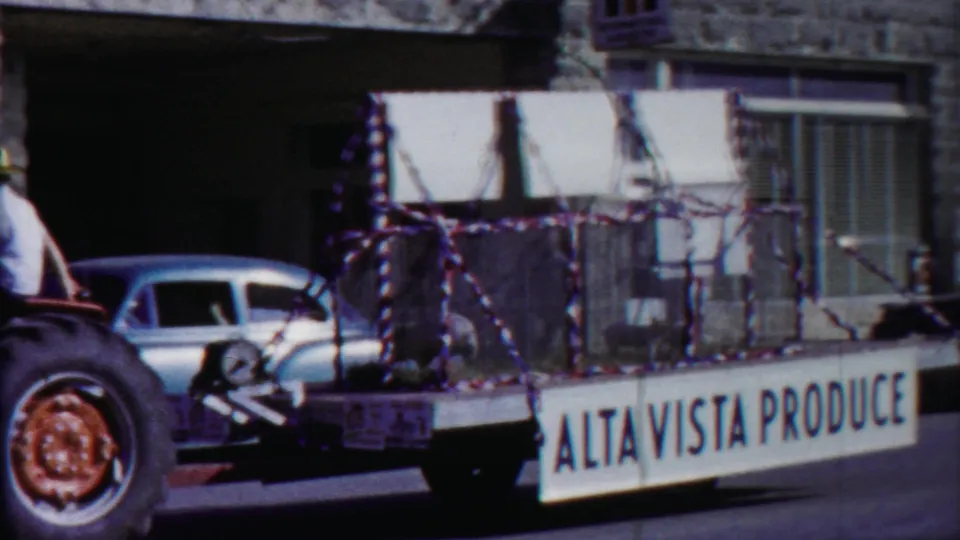 A trailer marked Alta Vista Produce appears beside the antique car, with parked sedans and a building facade in the background.