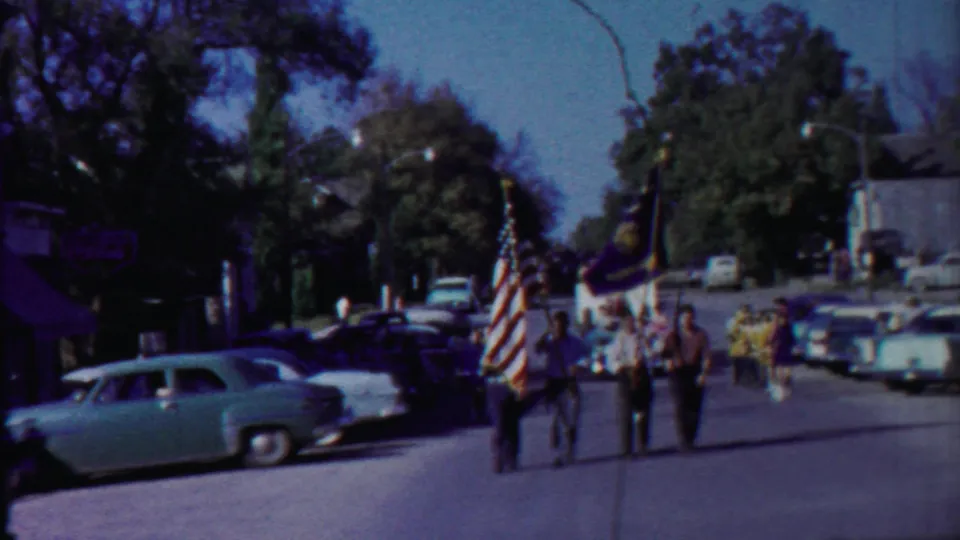 Men march in a 1958 Cheyenne County street parade with the American flag and other banners, with parked cars along the road.