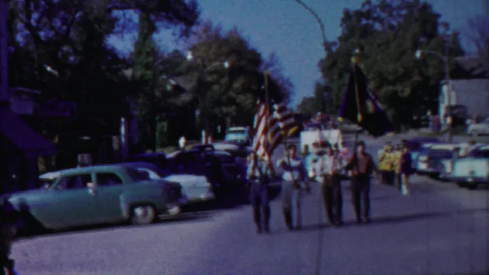 Cheyenne County parade participants carry flags along the roadway as classic cars and storefronts frame the small-town scene.
