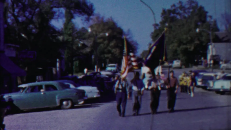 Three flag bearers in a 1958 Cheyenne County parade walk toward the camera, with spectators, parked cars, and trees behind them.