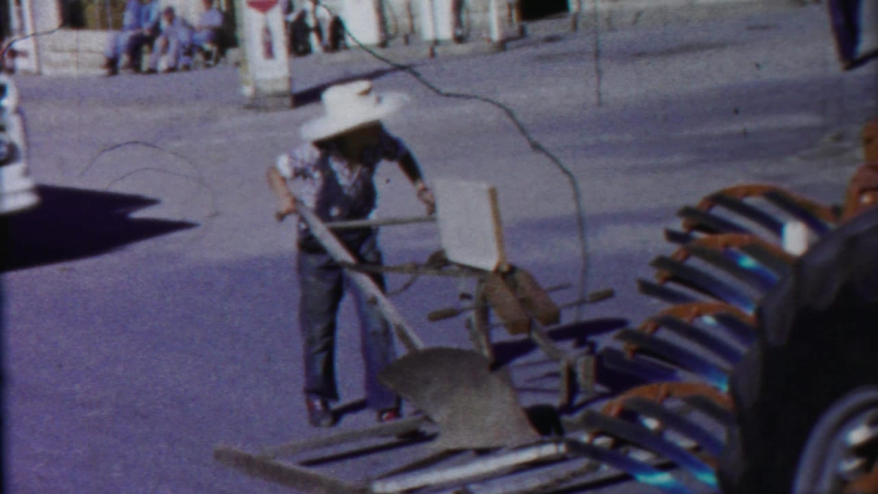 Man in a white hat pulling a farm implement across a paved street.