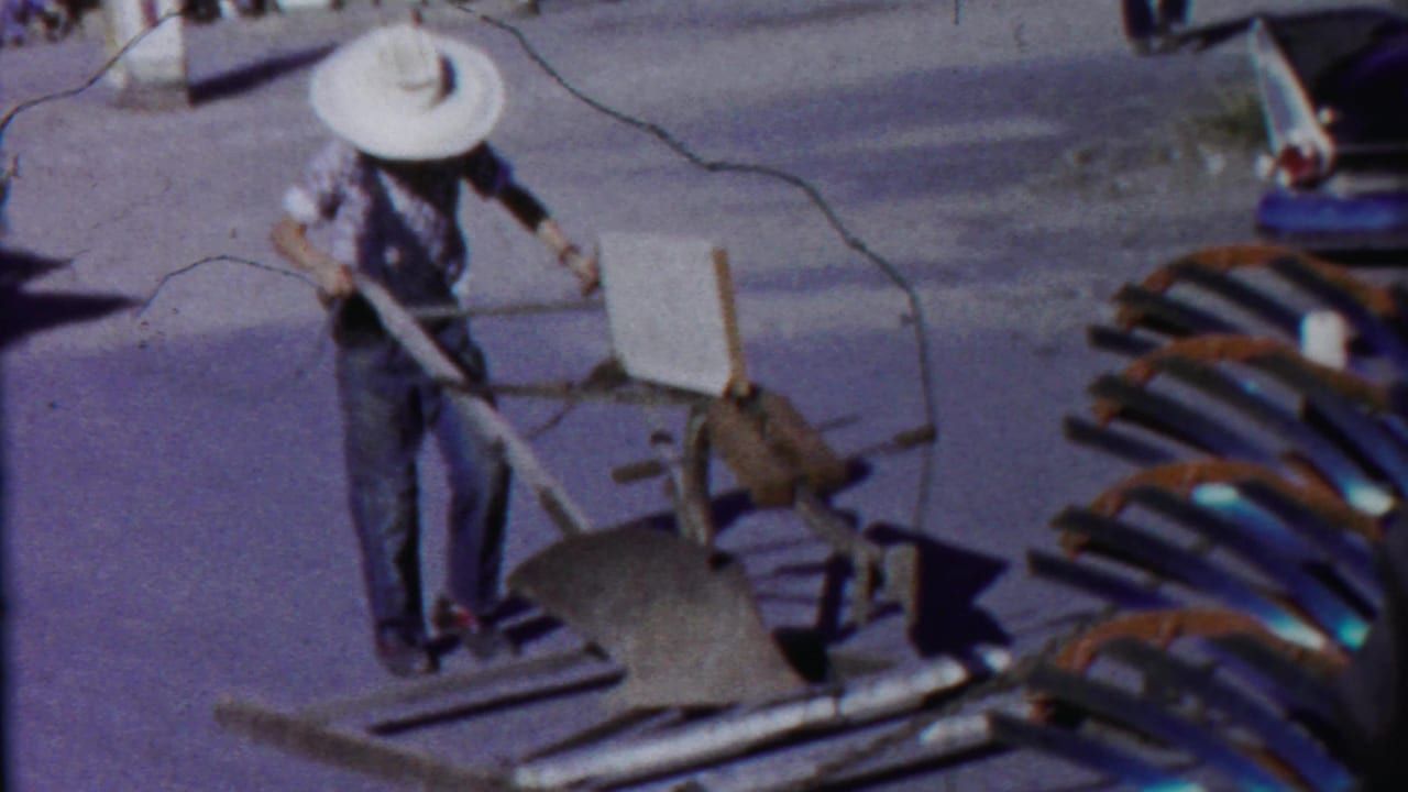 Man with a white cowboy hat guiding a wheeled farm implement on the street.