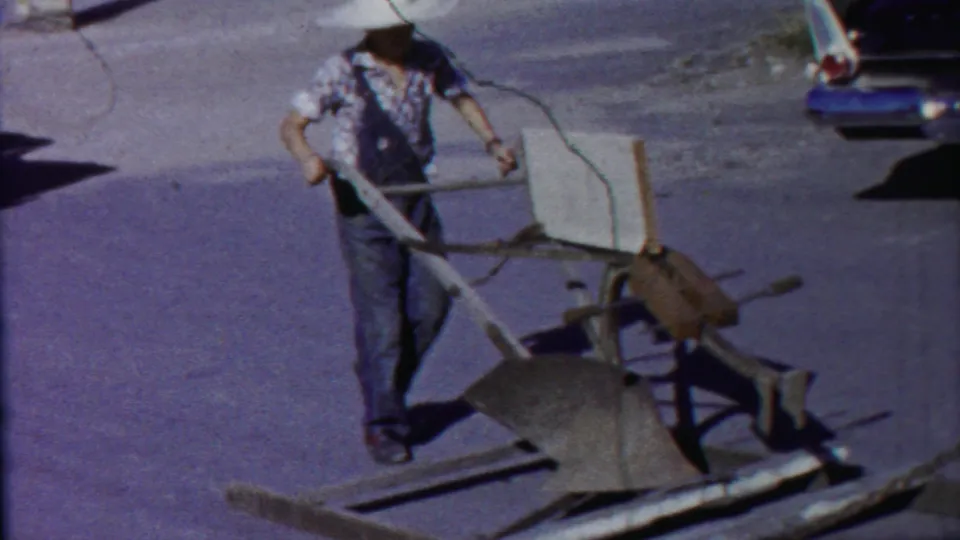 A man in work clothes pulls a plow-like implement along the pavement, with an older car and street shadows behind him.