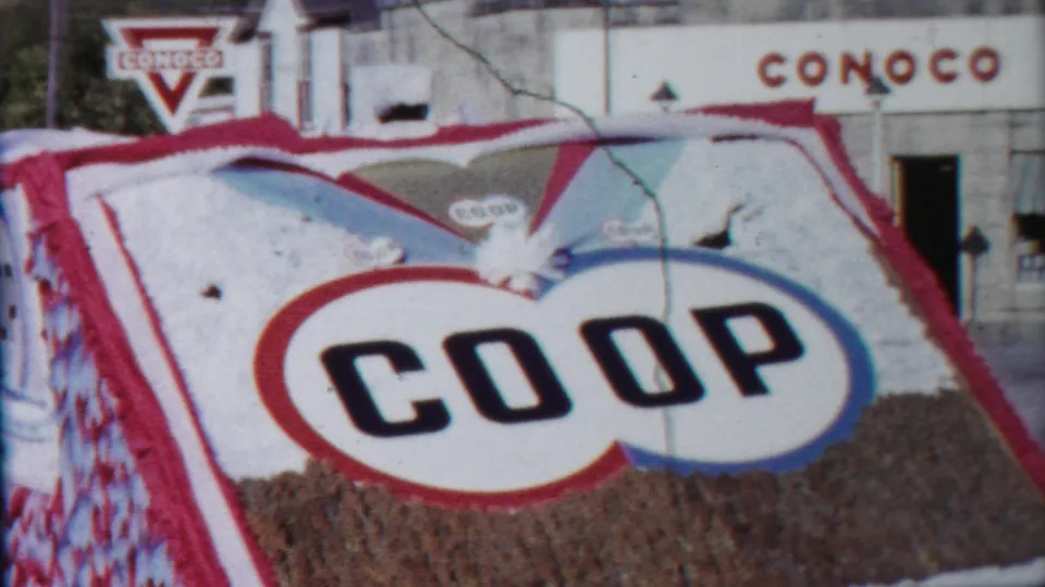 Handheld 8mm frame of the COOP float angled before a Conoco station, with a partial adult figure near the edge.
