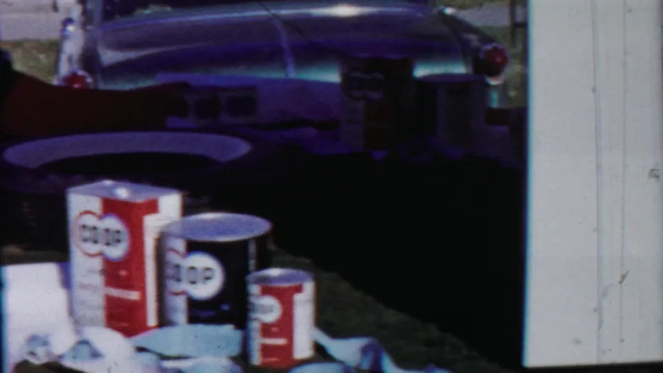 Outdoor display of COOP cans and boxed goods on grass, with a classic car bumper visible behind them in 1958 Kansas footage.