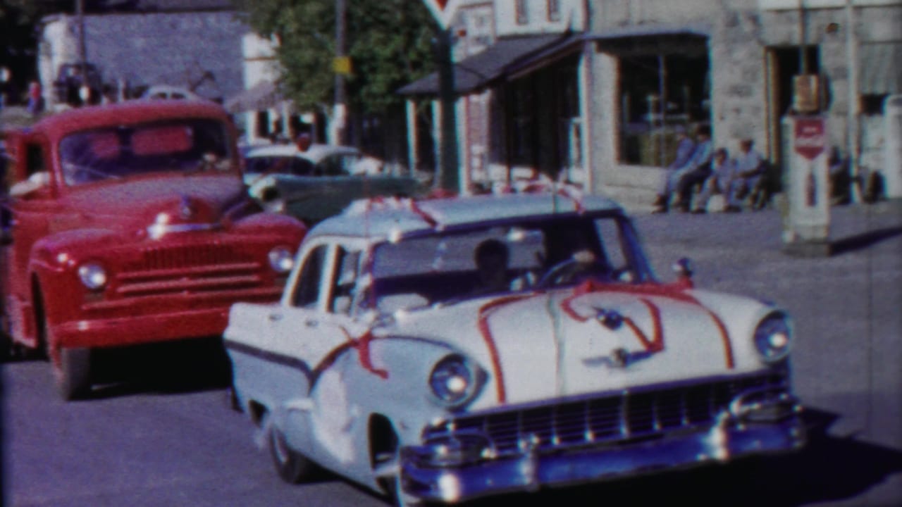 Ribboned white sedan passes a red truck on a small-town commercial street.