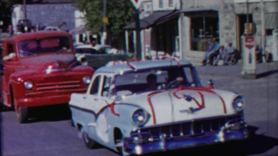 A decorated white 1950s sedan drives past a red truck on a Cheyenne County, Kansas street with storefronts and pedestrians nearby.