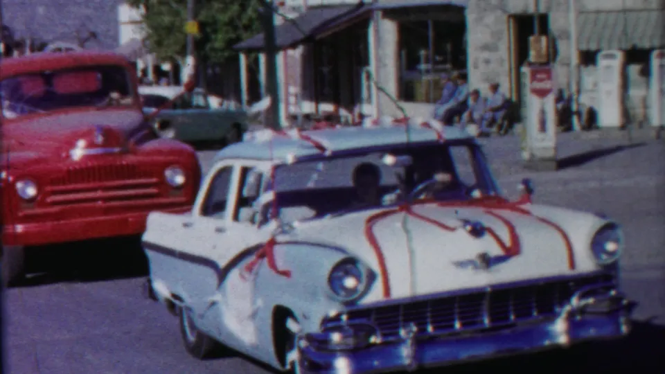 The ribboned wedding car moves close to the camera, with a red truck, gas pump, storefront windows, and seated people in the background.
