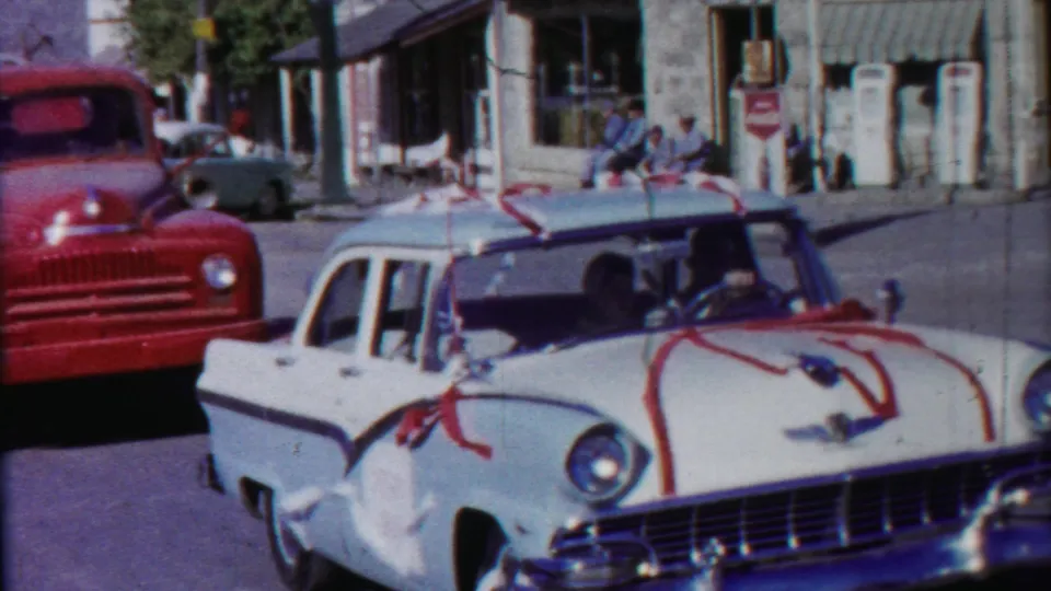 A white wedding-decorated sedan crosses the frame on a 1958 Kansas town street, with a red truck and seated pedestrians behind it.