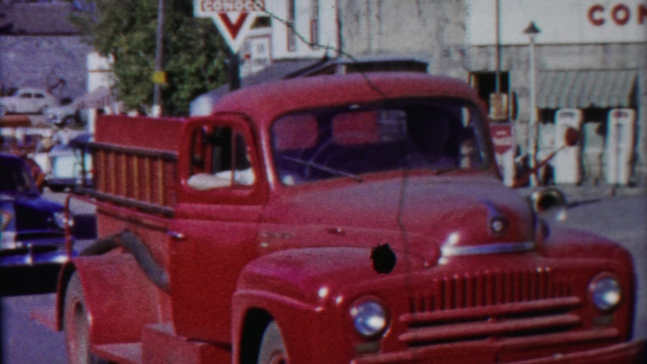 Red vintage fire truck with ladder parked on a small-town street near cars and storefronts.