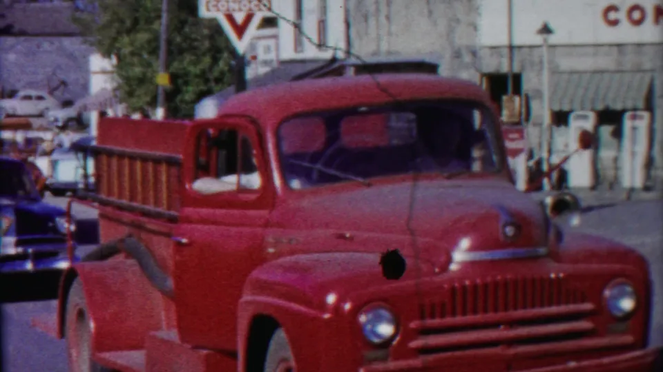 A red 1950s ladder truck is parked near a gas station and storefronts in Cheyenne County, Kansas, with period cars nearby.