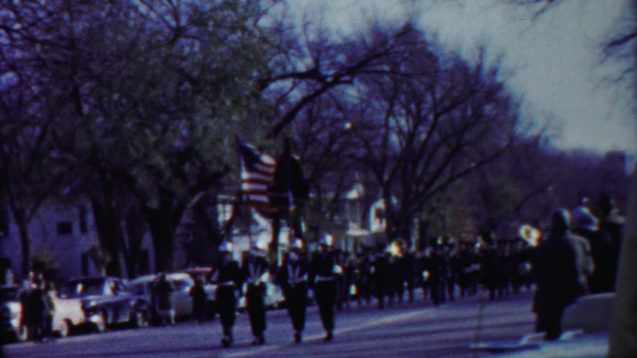 Band members and flag bearers march toward the camera past spectators and parked cars.