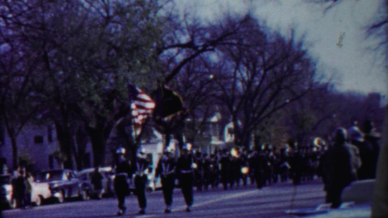 Mid-century Kansas parade footage shows uniformed band members approaching with flags as people watch from the curb.
