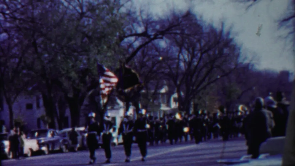 Mid-century Kansas parade footage shows uniformed band members approaching with flags as people watch from the curb.