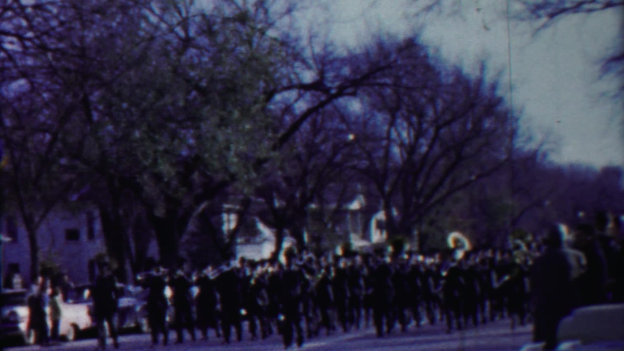 A marching band in dark uniforms advances through Cheyenne County, Kansas, with flag bearers and spectators visible in soft 8mm color.