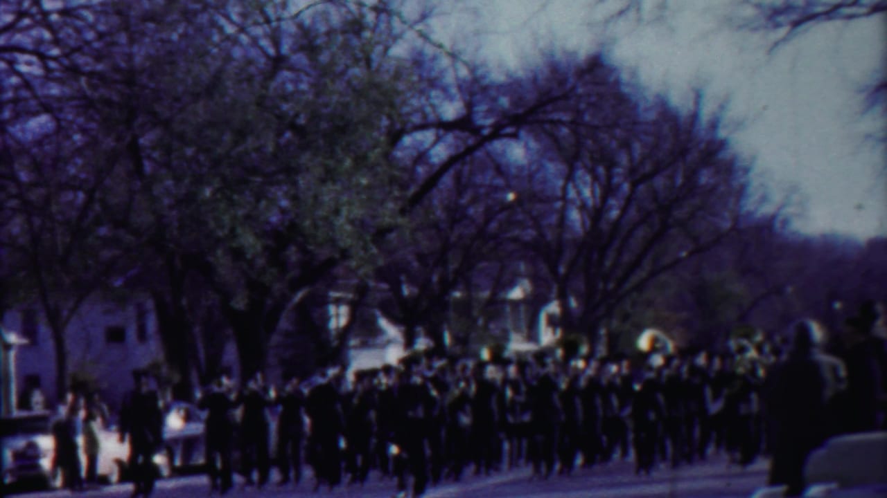 Marching band approaches along a tree-lined street with spectators nearby.