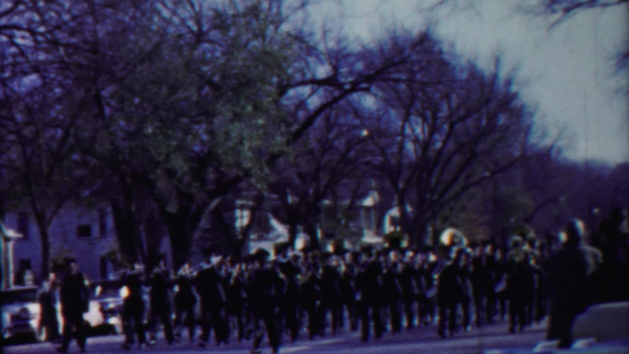 Marching band in dark uniforms continues along a Cheyenne County street, with a tuba visible above the group.