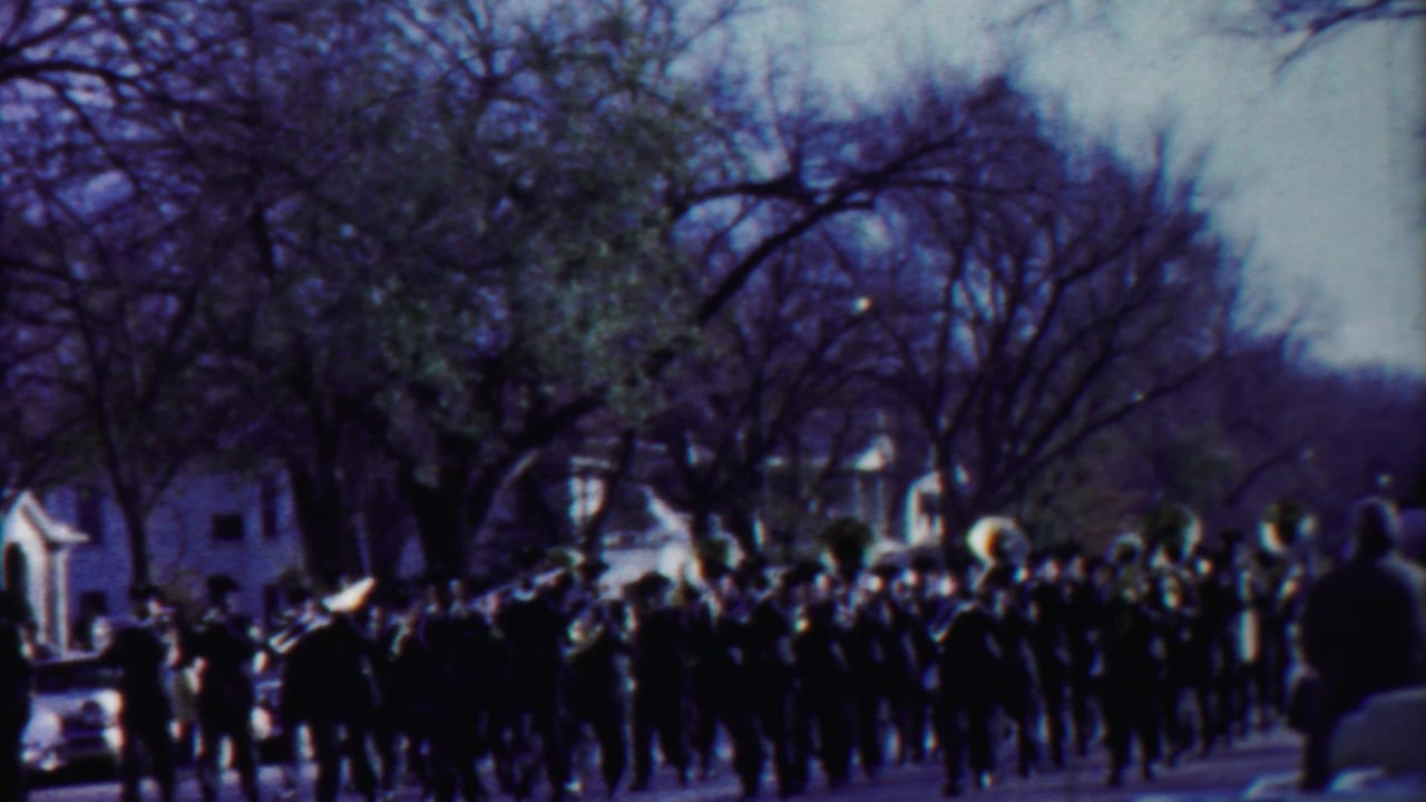 Marching band moves along a tree-lined street beside parked cars and a decorated float.