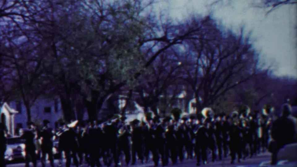 1958 Kansas home-movie frame showing a marching band on a residential street near a parade float and parked midcentury cars.
