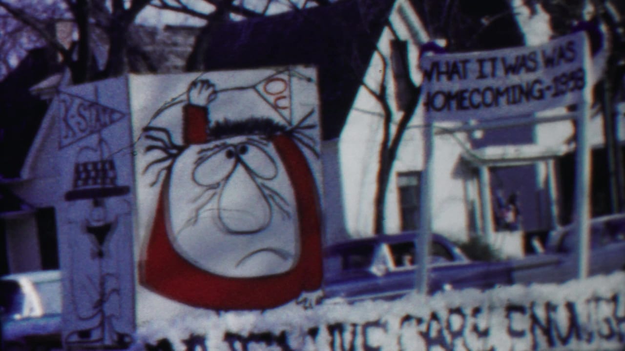 Kansas homecoming parade float passes residential houses, with a banner, cartoon artwork, and midcentury parked cars visible.