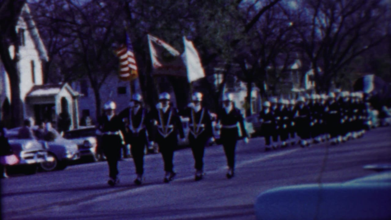 Uniformed marchers carry flags along a residential street with parked cars and houses.