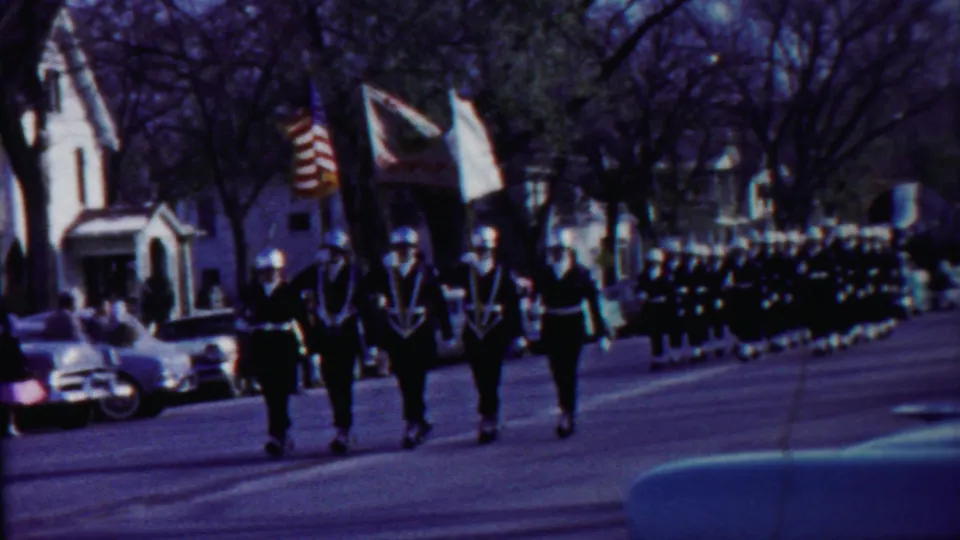 1958 Cheyenne County, Kansas home-movie frame showing uniformed marchers with flags moving down a residential street.