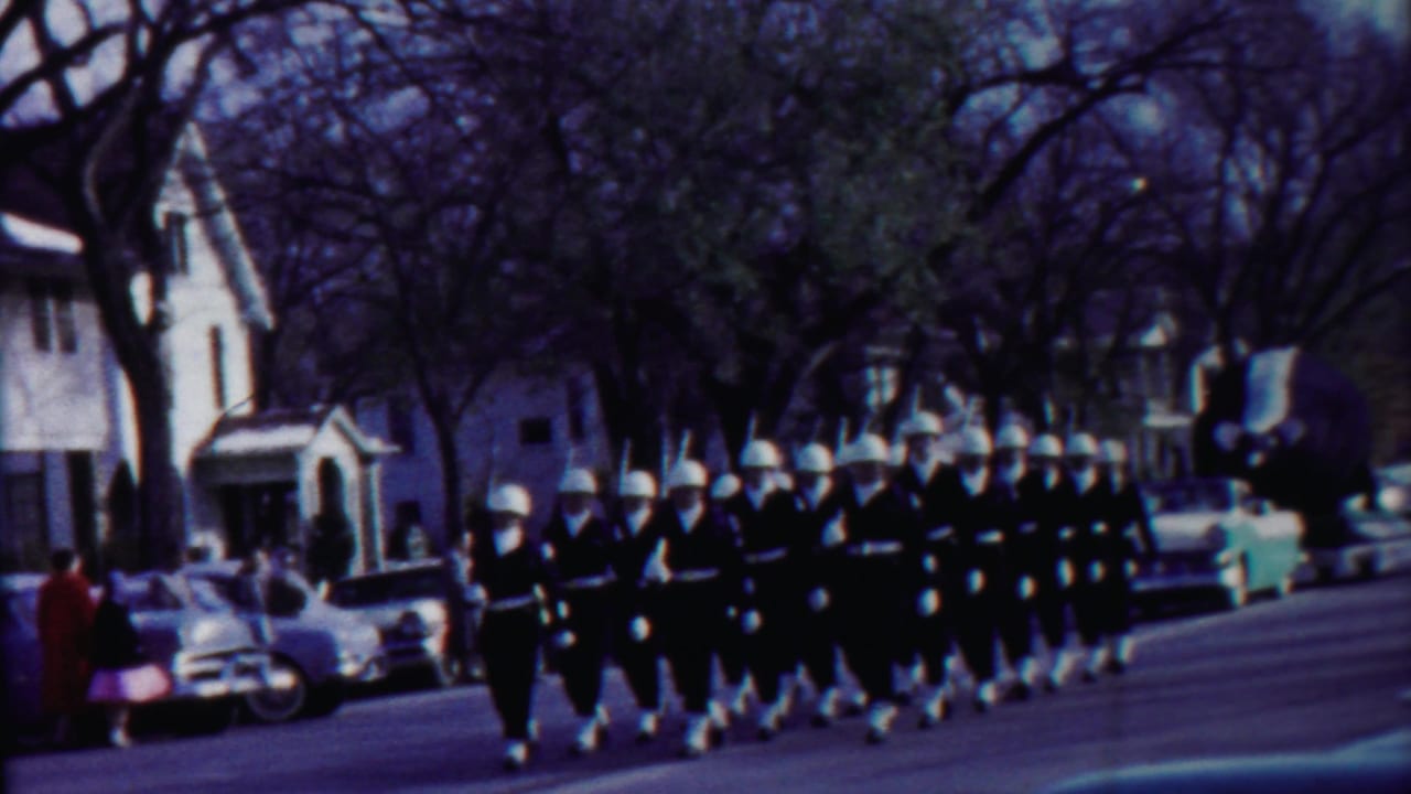 Helmeted uniformed marchers proceed in formation on a Cheyenne County residential street in 1958 footage.