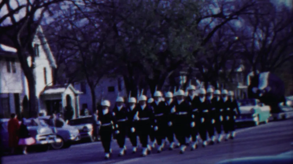 Helmeted uniformed marchers proceed in formation on a Cheyenne County residential street in 1958 footage.