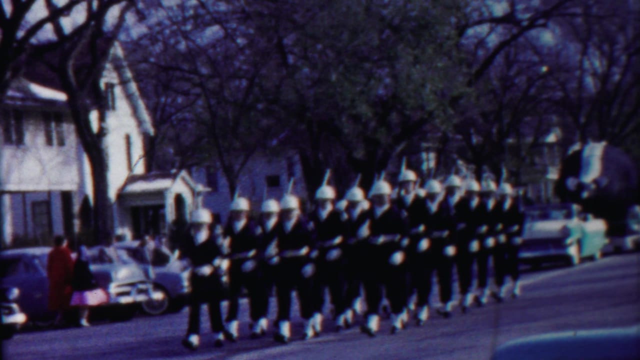 A formation of uniformed marchers passes houses, spectators, and mid-century cars in 1958 Kansas home-movie footage.