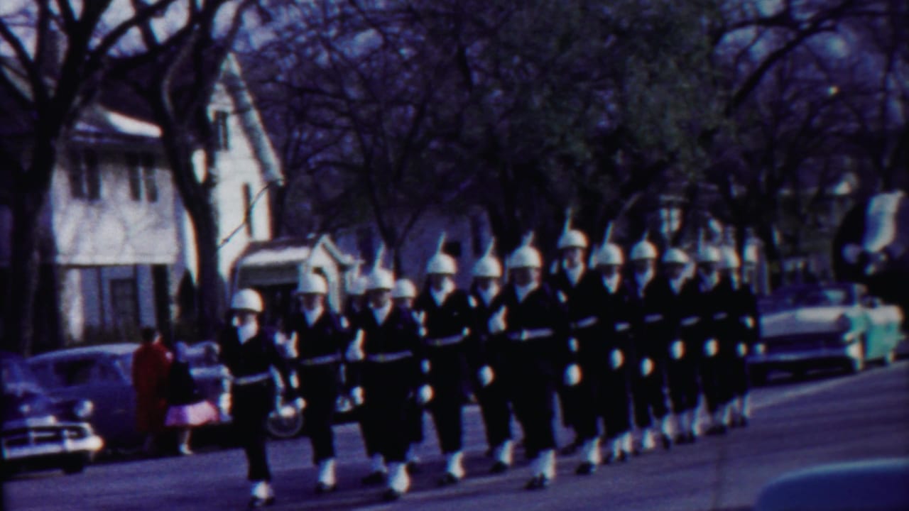 Close view of uniformed marchers in white helmets moving past a spectator and parked cars in Cheyenne County, Kansas.