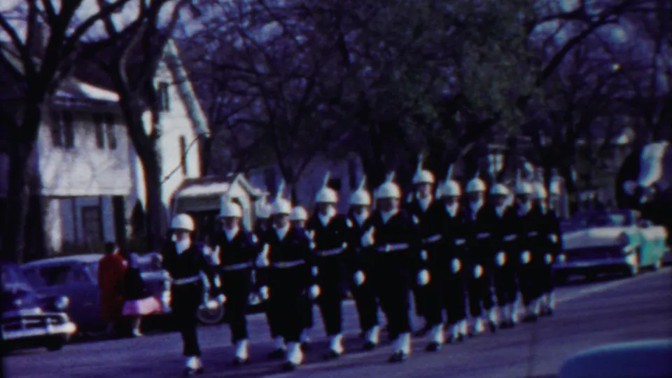 Close view of uniformed marchers in white helmets moving past a spectator and parked cars in Cheyenne County, Kansas.