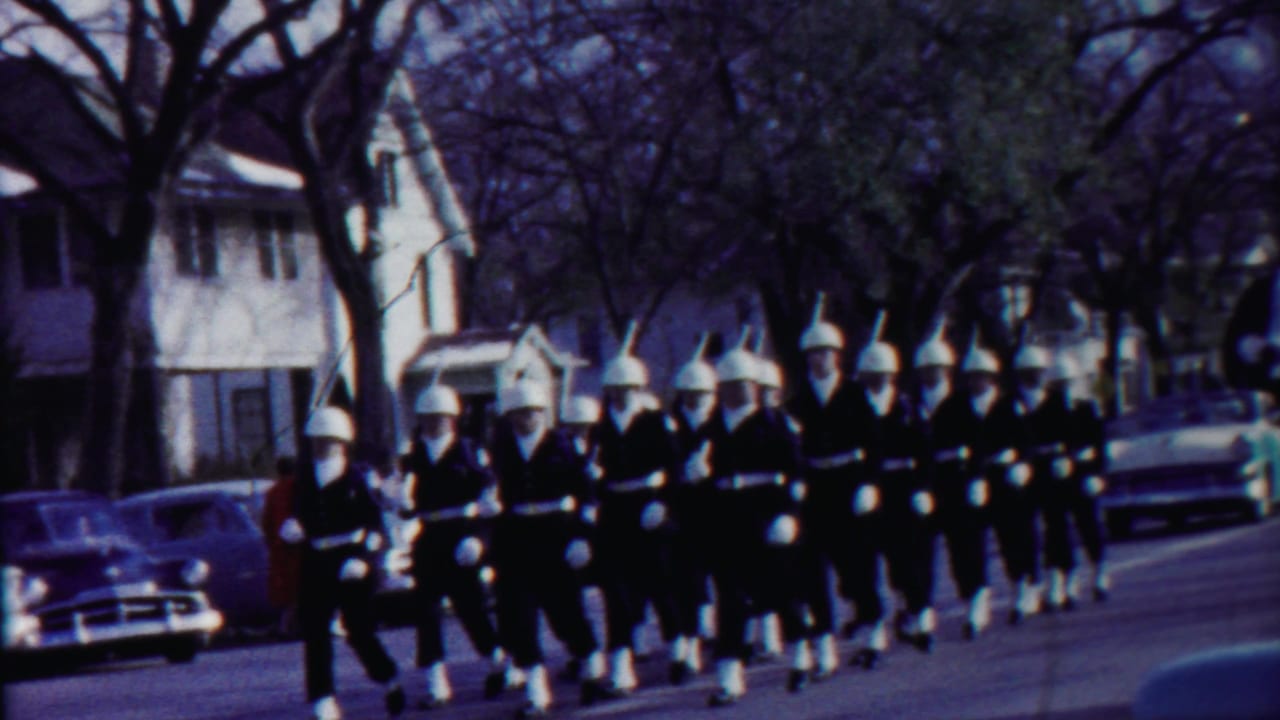 Marching band in white helmets moves along a residential street with parked cars and houses.
