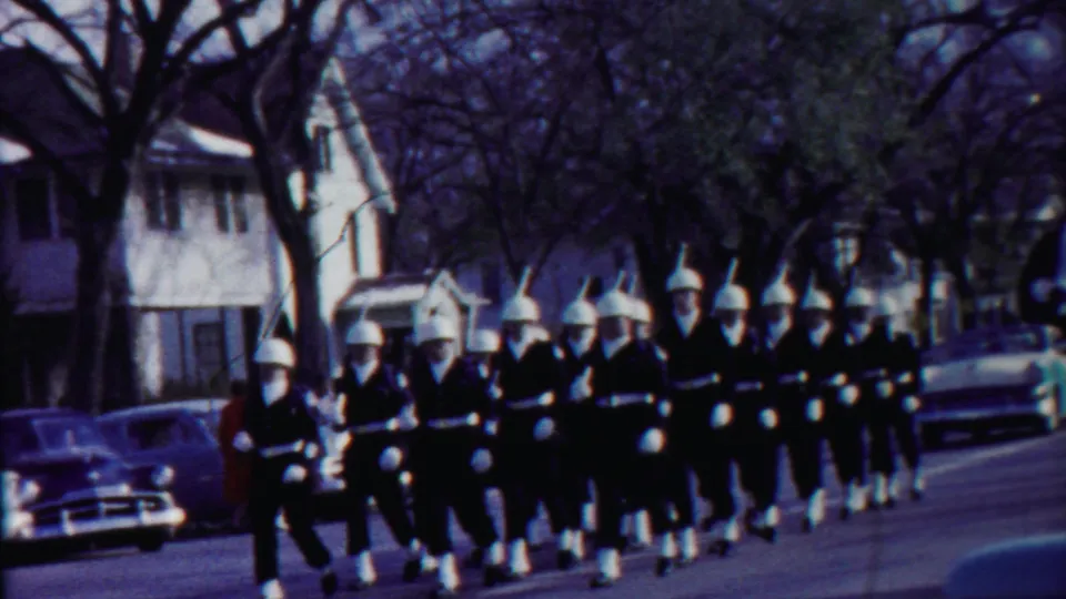 1958 Cheyenne County, Kansas home-movie frame showing a marching band in dark uniforms and white helmets on a neighborhood street.