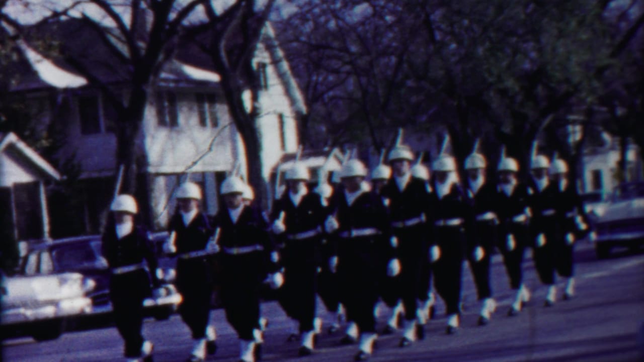 Curbside 8mm view of uniformed parade marchers in Cheyenne County, Kansas, passing houses and cars in 1958.