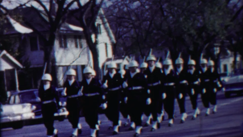 Curbside 8mm view of uniformed parade marchers in Cheyenne County, Kansas, passing houses and cars in 1958.