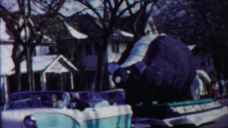 A large bear-shaped parade float moves through a Cheyenne County, Kansas residential street in 1958 home-movie footage.