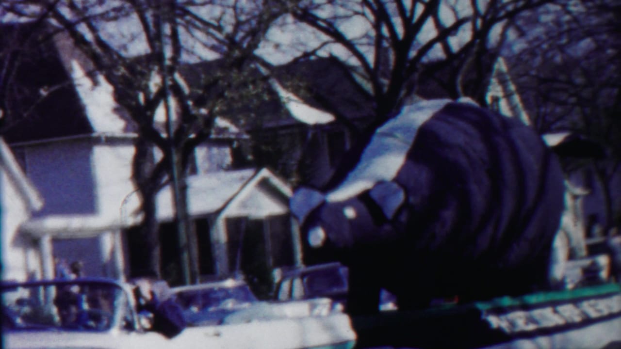 The bear float is seen from the side as it passes homes and parked cars during the 1958 Kansas parade footage.