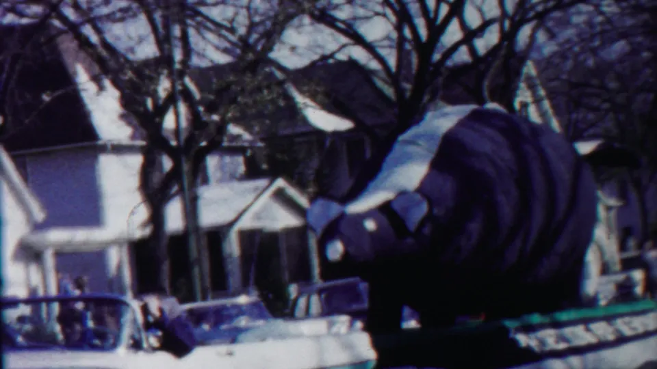 The bear float is seen from the side as it passes homes and parked cars during the 1958 Kansas parade footage.