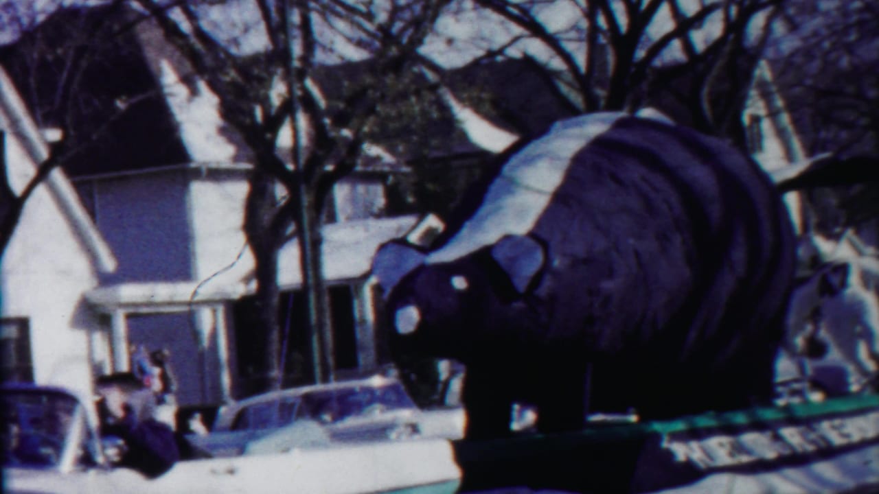 Giant skunk parade float passing houses and leafless trees with onlookers nearby.