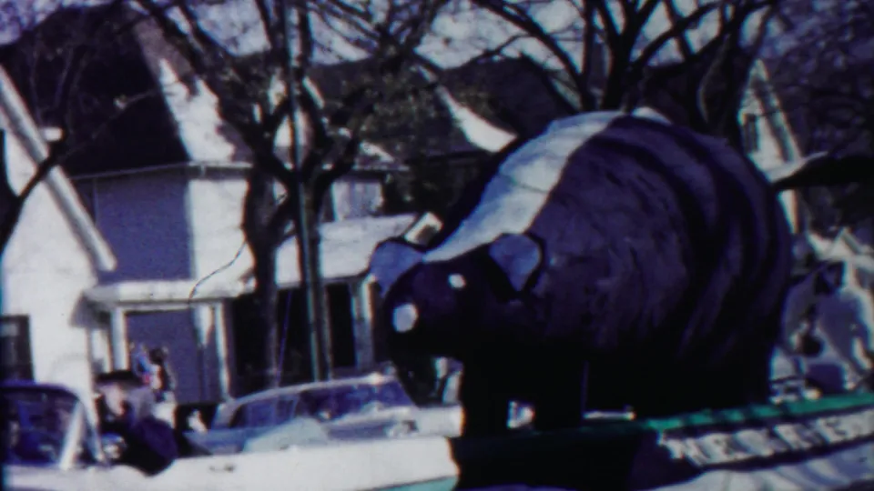 In 1958 Cheyenne County, Kansas footage, a large dark skunk float with a white stripe moves past houses, trees, cars, and spectators.