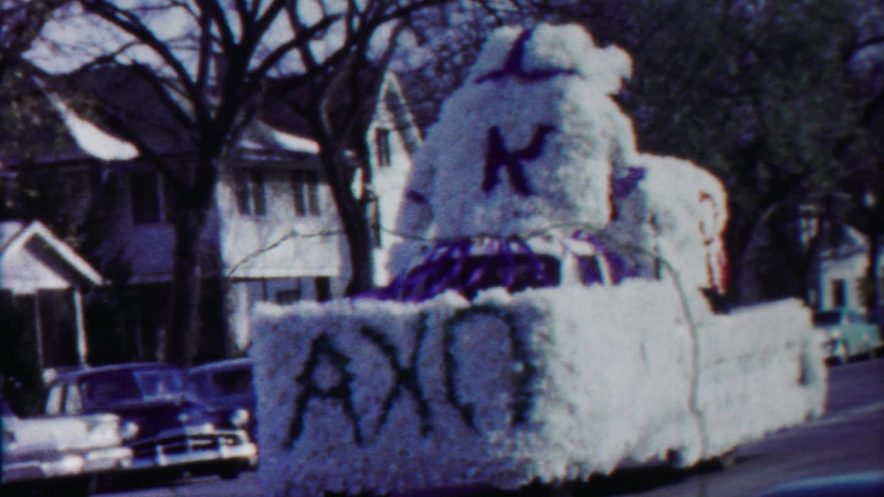 1958 Kansas home-movie frame of a white parade float with a large K and purple trim moving through a residential street.