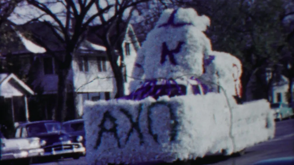 1958 Kansas home-movie frame of a white parade float with a large K and purple trim moving through a residential street.